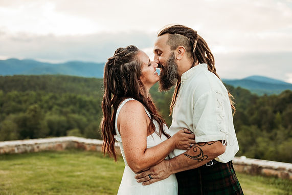A couple smiles at each other as they lean in for a kiss in front of a scenic mountain view.