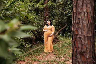 A pregnant woman wearing a yellow lace dress has hands on her bump while standing surrounded by trees.
