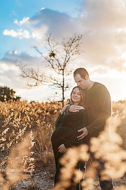 A pregnant mom leans against her husband as they embrace in a field.