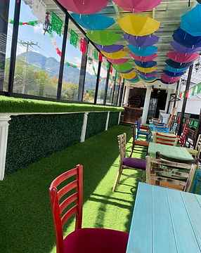 Terraza exterior con sombrillas coloridas, pasto artificial, sillas y vista a montañas.