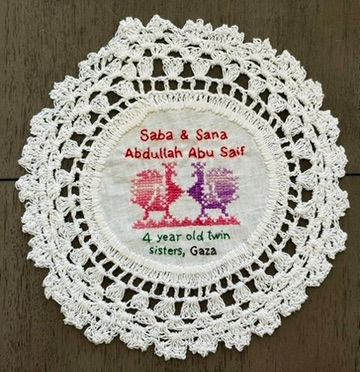 Tatreez pattern of two peacocks embroidered on a doily with the names of Saba and Sana Abdullah Abu Saif, 4 year old twin sisters from Gaza. The names have been embroidered in English.