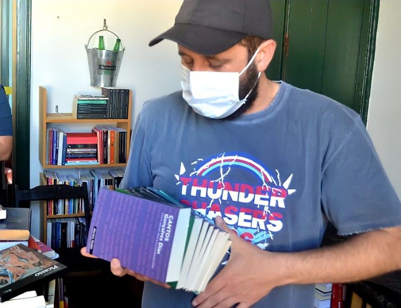 Candido está com máscara branca e camisa azul, segurando vários livros.