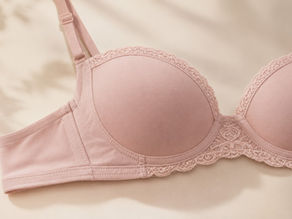 Close-up of a soft blush pink balconette bra with smooth molded cups and delicate lace trim, photographed in warm natural light on a cream fabric background.