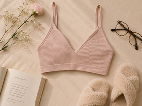 A lifestyle photograph of a soft pink ribbed wireless bralette resting on beige linen sheets next to glasses, an open book, and white carnations.