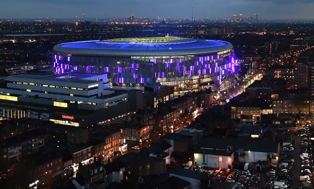 Illuminated stadium at night with blue and purple lights, surrounded by cityscape. Sainsbury's visible in foreground, creating a vibrant scene.