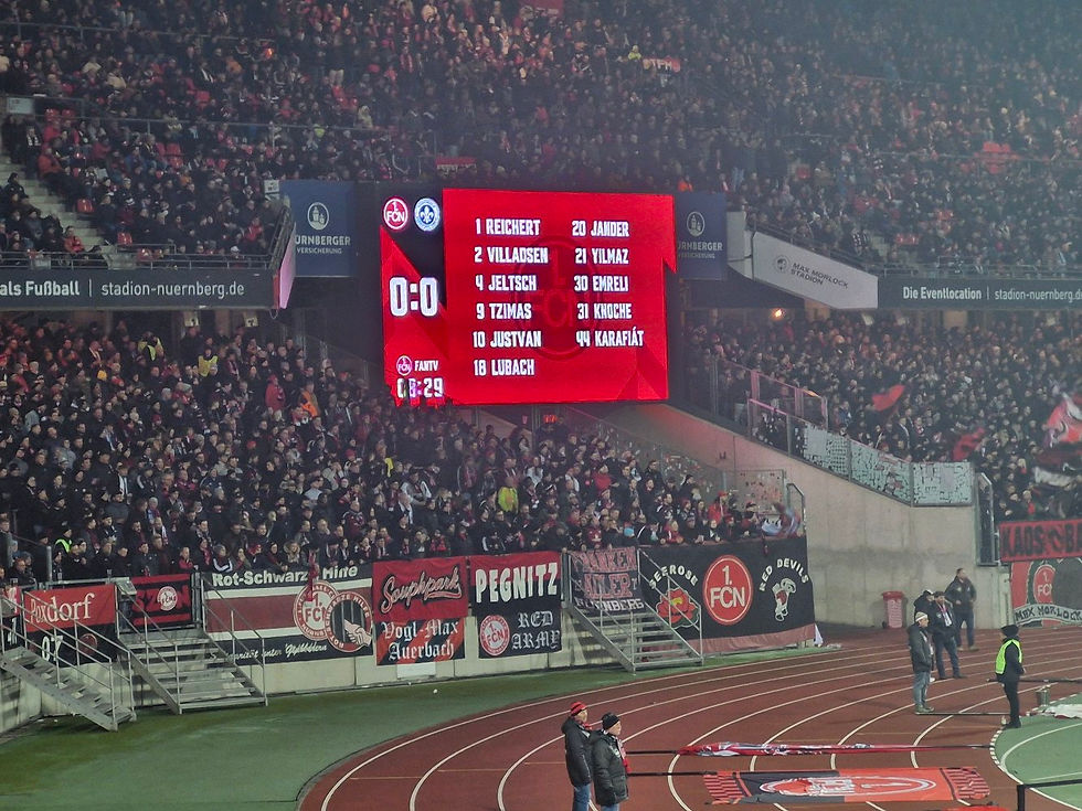 Packed soccer stadium with a red scoreboard showing 0:0 and team lineups, fans filling the stands in a tense atmosphere.