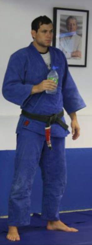 Man in a blue judo gi holds a water bottle while standing on a mat, with a framed portrait on the wall behind him.