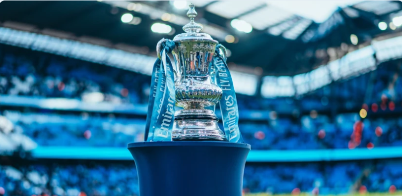 Silver trophy with blue ribbons on a stand in a stadium. Blue seating and partially cloudy sky visible, creating a celebratory atmosphere.