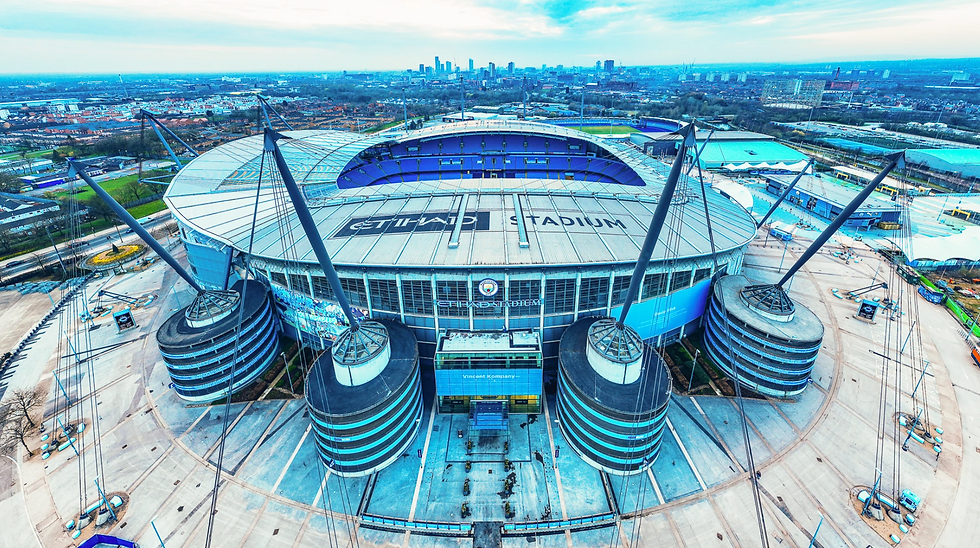 Aerial view of Etihad Stadium, Manchester, with blue seats, gray roof, and surrounding cityscape under a cloudy sky. Stadium and city names visible.