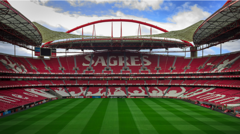 Empty red-and-white soccer stadium with green striped pitch and SAGRES seats under a blue sky.