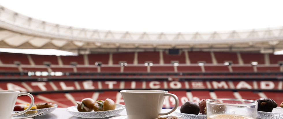 Coffee cups and pastries on a table overlooking a red stadium with blurred text "ATLETICO DE MADRID" on the seats. Bright and inviting.