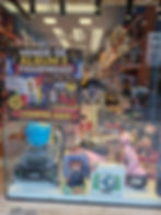 Toy store window with FIFA 2026 sticker album sign, dolls, Lego and ride-on scooter; bright shelves and colorful boxes.