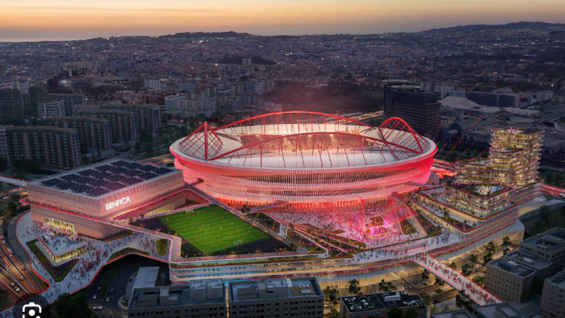 Aerial view of Benfica’s illuminated stadium complex at dusk, with red lights, city buildings, and visible Benfica text.