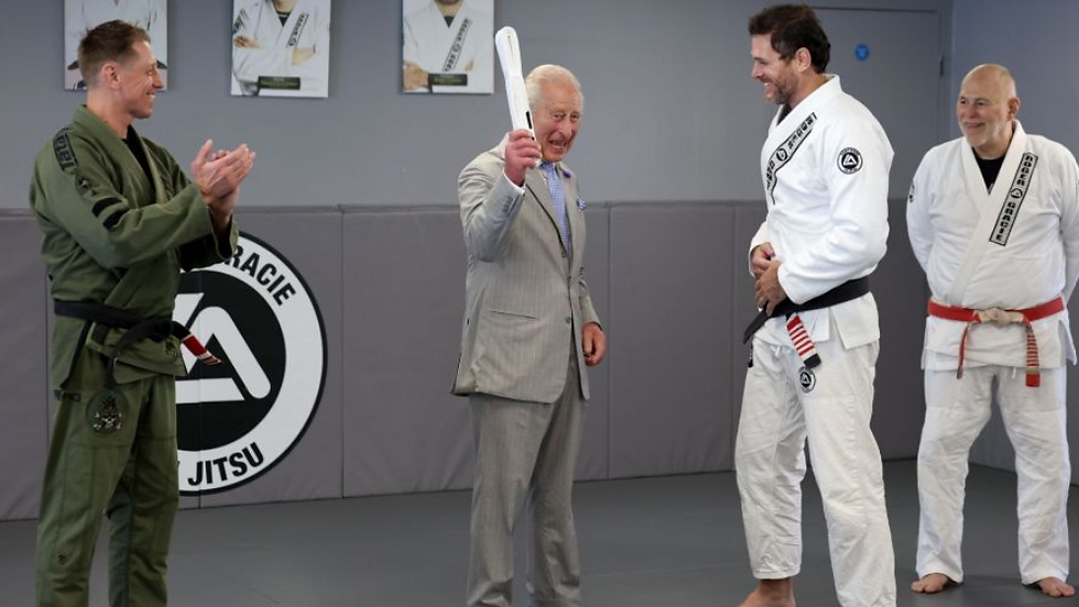 Elderly man in a suit raises a white belt in a jiu-jitsu dojo as three men in gis smile and applaud.