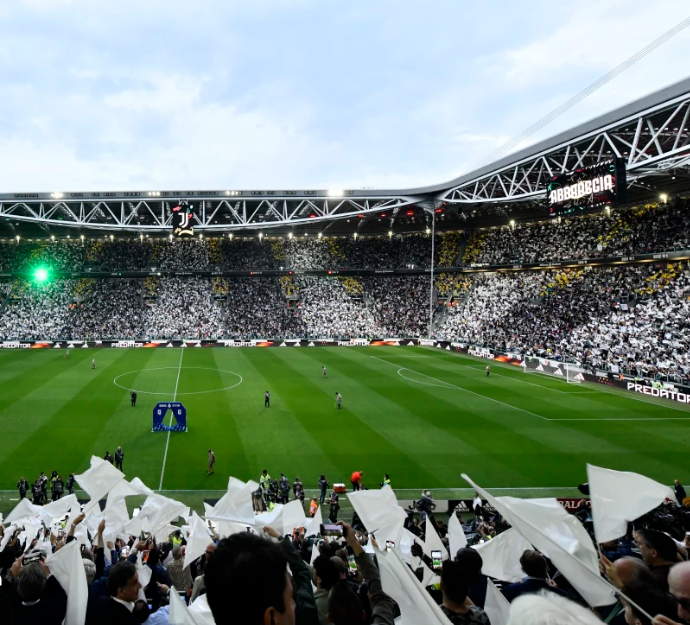 Packed soccer stadium with fans waving white flags around a green pitch; large crowd and scoreboard banner reading ABBODCIA.