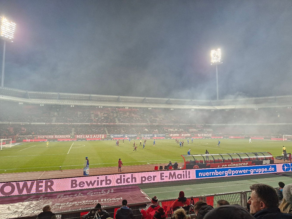 Foggy night soccer match in a packed stadium, players in red and blue on the field, with ads reading Bewirb dich per E-Mail and nuernberg@power-gruppe.de
