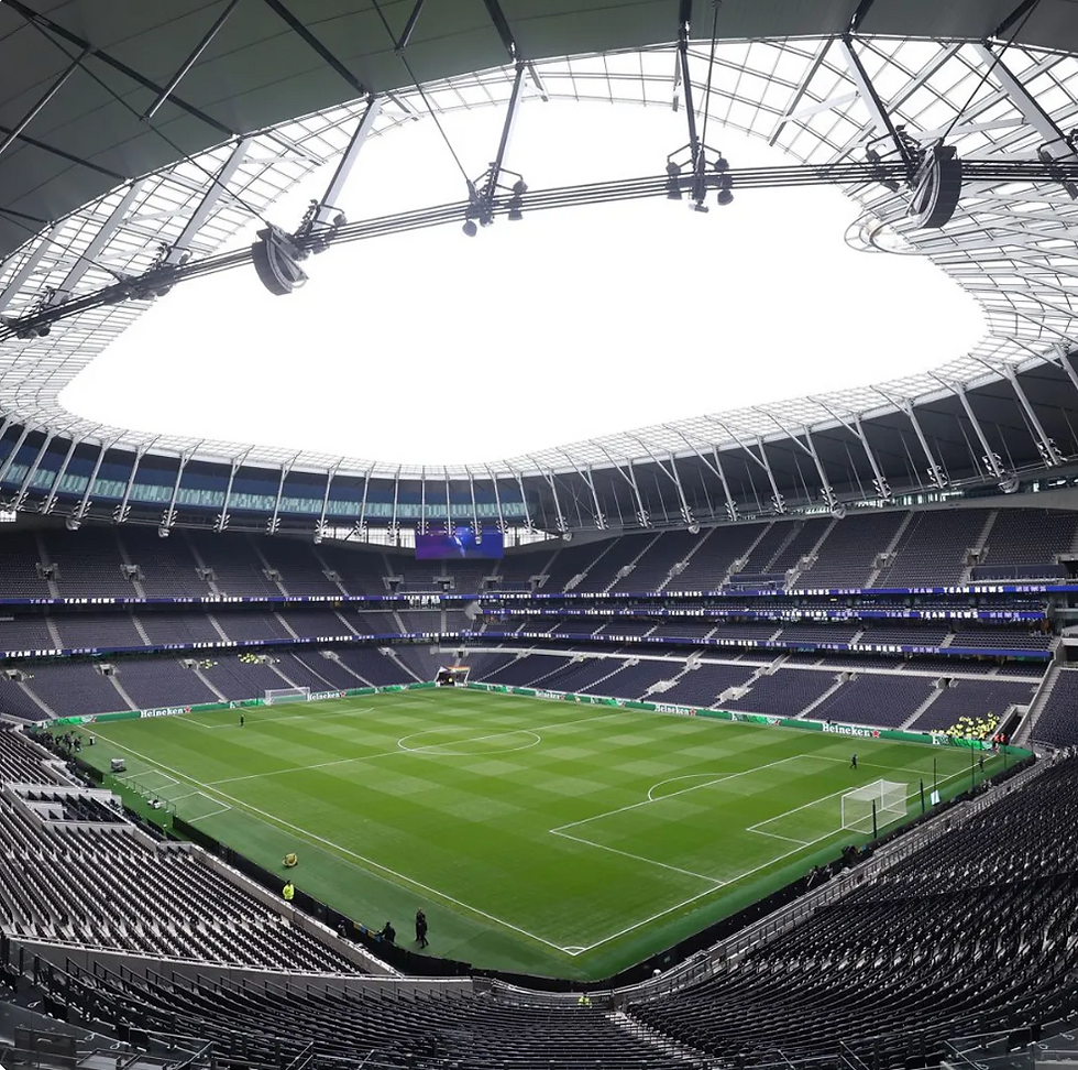 Empty modern stadium with green soccer field and rows of dark seats under a curved roof. Large screens display "TEAM NEWS." Quiet atmosphere.