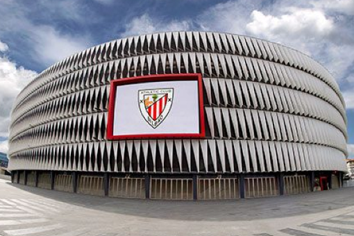 Modern stadium exterior with silver ribbed facade and red-framed Athletic Club crest under a cloudy sky.