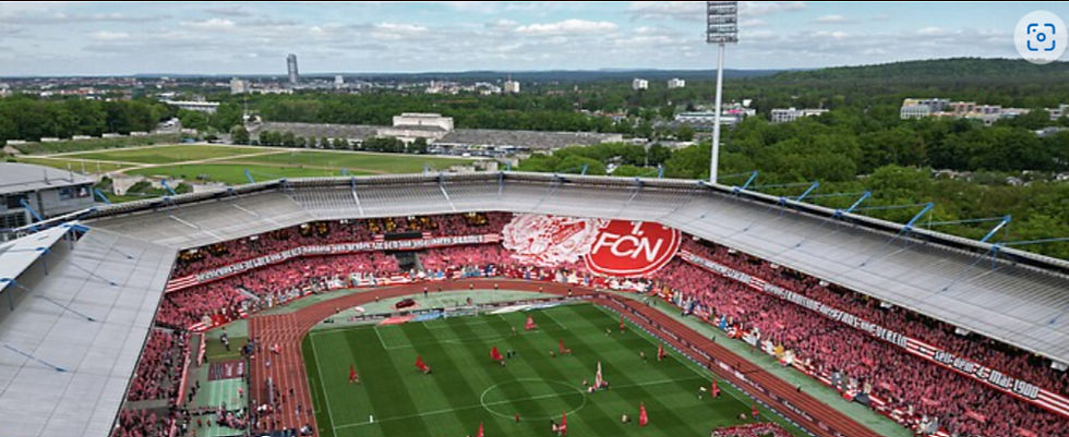 Aerial view of a packed soccer stadium, red-clad fans waving a huge FCN banner around the green pitch during a match.