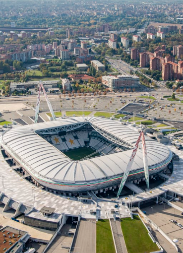 Aerial view of a large white-roofed stadium in a city, with surrounding roads, apartments, and green spaces under clear daylight.