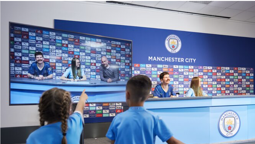 Children in blue jerseys point at a video screen showing a press conference at Manchester City. Bright backdrop with sponsor logos.