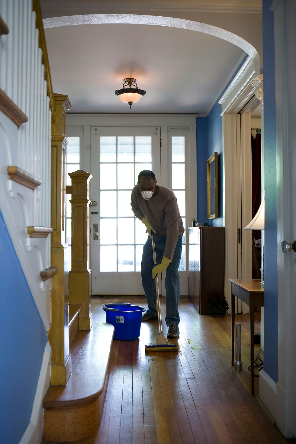 Person mopping wooden floor in a blue-walled room. Wearing mask and gloves, next to a blue bucket. Bright light from door creates a calm mood.