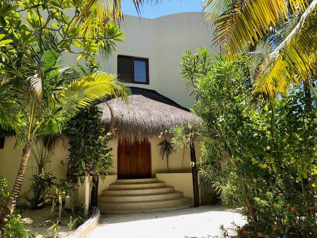 Casa Mariposa oceanfront home in Tankah Bay, Tulum, with tropical gardens, white facade, and a palapa-covered entrance.