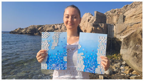 Woman smiles at a rocky beach, holding two blue abstract paintings with mosaic-like white patterns.