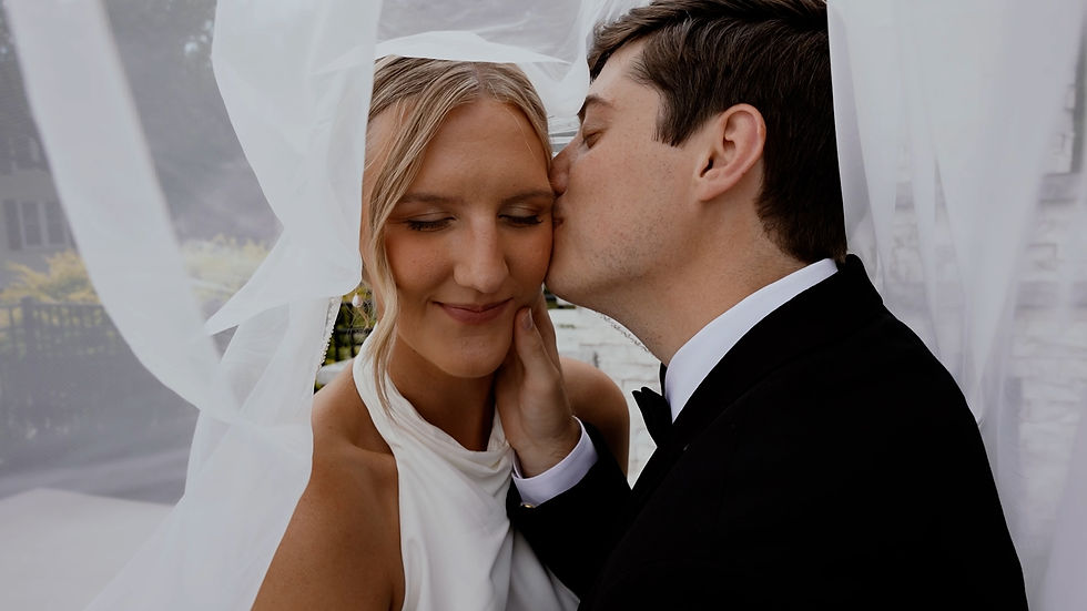 Groom kisses smiling bride on the cheek under white wedding veil, intimate romantic close-up outdoors