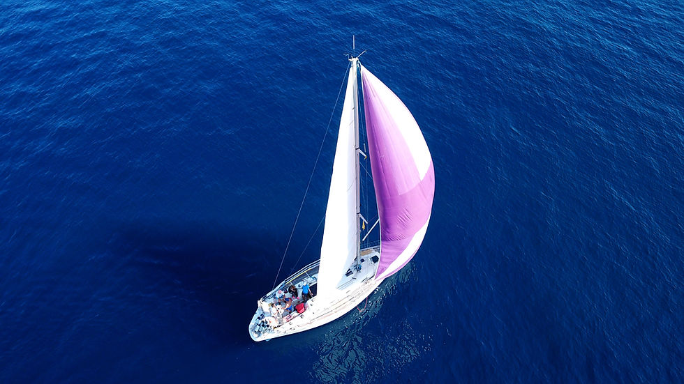 Aerial view of a yacht sailing in deep blue sea, with a crew on deck. It has a large white sail and a vibrant purple spinnaker.