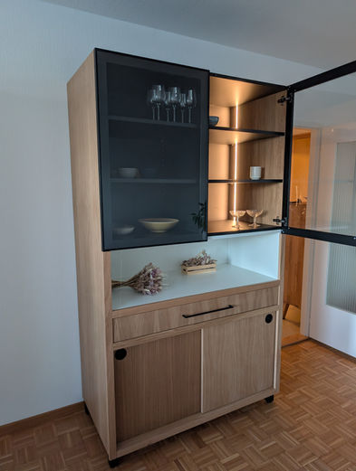 Vaisselier sur mesure en chêne avec portes coulissantes et vitrines en verre transparent sur cadre métallique noir. Meuble de rangement haut de gamme avec éclairage LED intégré, idéal pour salle à manger ou séjour. Fabrication artisanale par une menuiserie à Martigny en Valais, spécialisée dans le mobilier sur mesure, alliant design contemporain, matériaux nobles et finitions soignées.