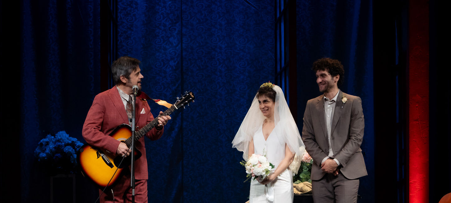 nella foto sulla sinistra c'è un uomo in completo rosso mattone che suona la chitarra, sulla destra una coppia di sposi, lei in bianco con il boquet in mano, lui con un abito grigio è sorridete alla sua destra. uno spettacolo che andrà in scena al Teatro Bobbio de La Contrada di Trieste