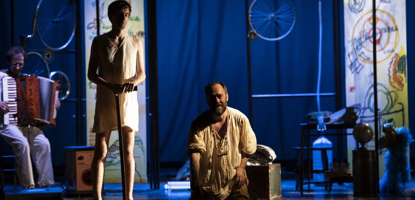 Archimede, la Solitudine di un genio, è lo spettacolo che andrà in scena presso la sala Bobbio del teatro La Contrada. L'attore Mario Incudine ah centro inginocchiato, indossa una camicia morbida beije e collane lunghe al collo. Sguardo verso lo spettatore. Background blu, uomo in piedi alla sua sinistra e in ultimo piano un musicista seduto con una fisarmonica.