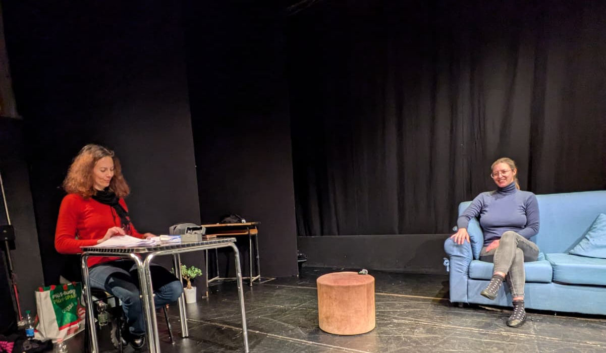 Pizza Burek è la residenza artistica che andrà in scena presso il teatro dei Fabbri di Trieste. Attrice a sinistra con maglioncino rosso. Sulla destra infondo, un divano azzurro con una signora seduta a gambe incrociate. Esse si trovano sul palcoscenico.