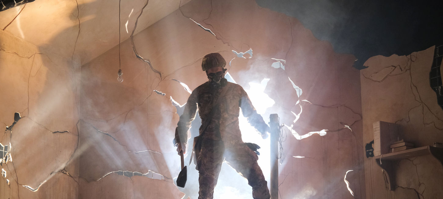 Un uomo in mezzo a una stanza con delle crepe al muro. Le pareti sono rosa aranciate e si intravede la luce nelle crepe. Egli indossa abiti militari, visibile solo la sua sagoma controluce. Egli è in piedi su un letto con delle coperte. Luci soffuse. The Other Side è uno spettacolo che andrà in scena al Teatro Bobbio de La Contrada di Trieste.