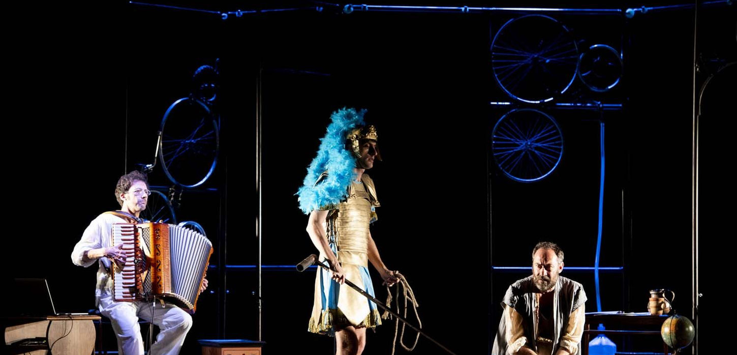 Archimede, la Solitudine di un genio, è lo spettacolo che andrà in scena presso la sala Bobbio del teatro La Contrada. L'attore Mario Incudine ah centro inginocchiato, indossa una camicia morbida beije e collane lunghe al collo. Sguardo verso lo spettatore. Soldato sulla sinistra in piedi, indossa un elmo con piumaggio azzurro. In ultimo piano un musicista seduto con una fisarmonica. Sfondo blu.