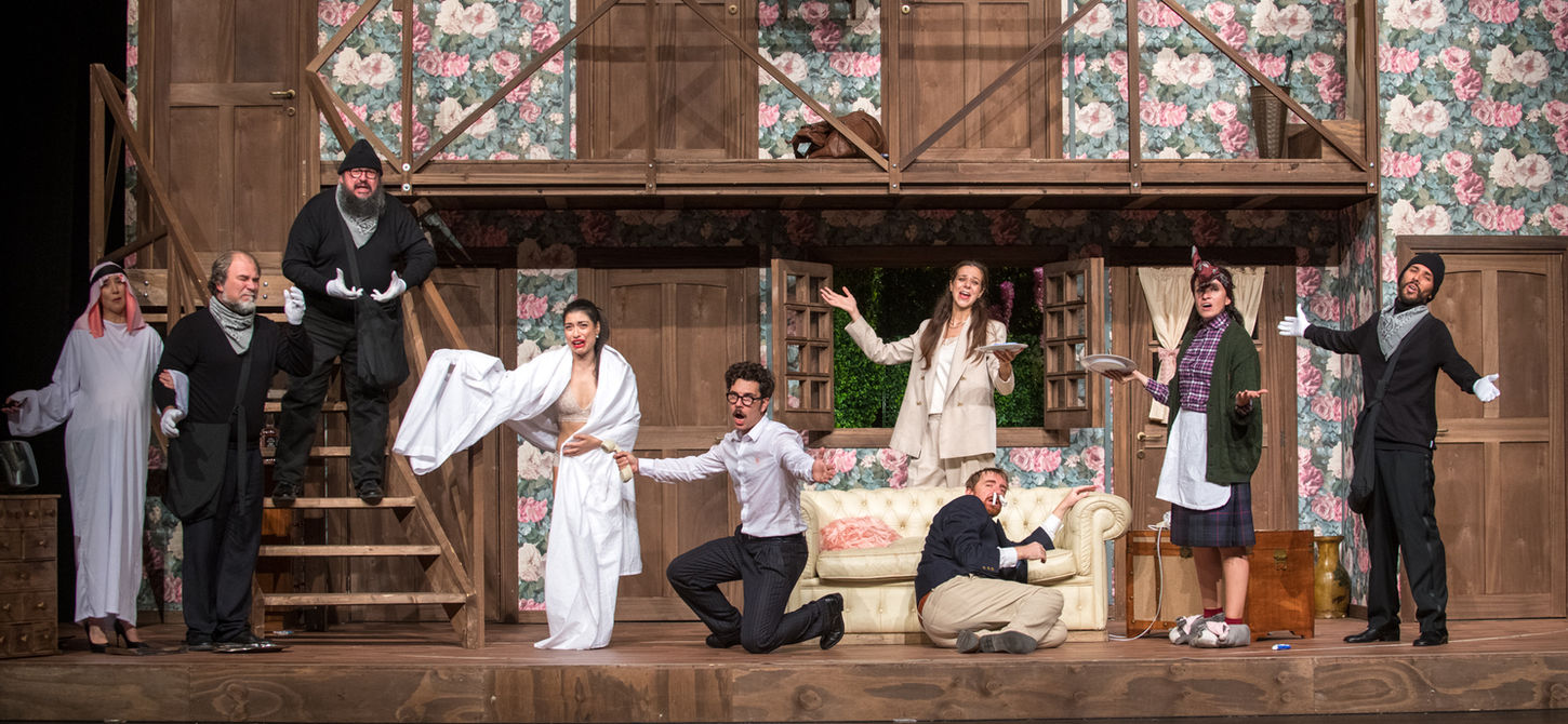 la foto di scena di "Rumori Fuori Scena", gli attori distribuiti sul palco hanno le braccia alzate. Lo sfondo è composto dal salotto, le scale, al secondo piano ci sono 3 porte. uno spettacolo che andrà in scena al Teatro Bobbio de La Contrada di Trieste
