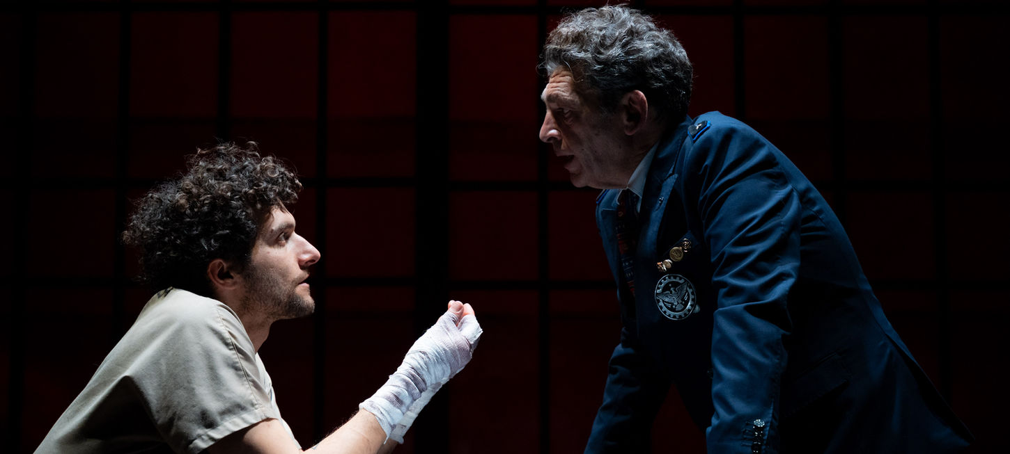 Nella foto un attore di profilo è seduto e appoggia il gomito sul tavolo, con il braccio verso l'uomo di fronten a lui, ha il braccio fasciato. l'uomo di fornte a lui, anche lui di profilo ha il corpo un po' piegato verso il ragazzo. Lo sfondo è nero. uno spettacolo che andrà in scena al Teatro Bobbio de La Contrada di Trieste