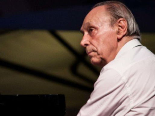 Livio Cecchelin in primo piano con camicia bianca, seduto al pianoforte. Girato in obliquo con sguardo verso sinistra. Volto con espressione seria e bocca serrata.