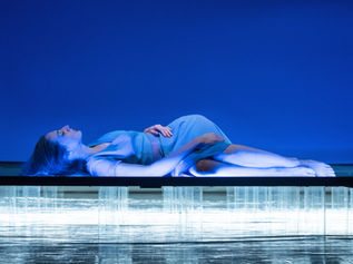 Anna Della rosa stesa con gambe raggomitolate su e stessa. Mano sul ventre, volto rivolto verso il cielo con occhi chiusi. Luci soffuse blu. Spettacolo per la stagione teatrale de La Contrada.