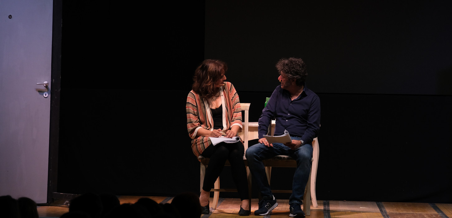 Marzia Postogna e Maurizio Repetto per lo Spettacolo La Felicità. Entrambi seduti su delle sedie in legno. Si stanno guardando. lei indossa uno scialle a quadri, lui una giacchetta blu. Background nero. Uno spettacolo che andrà in scena al Teatro Bobbio de La Contrada di Trieste.