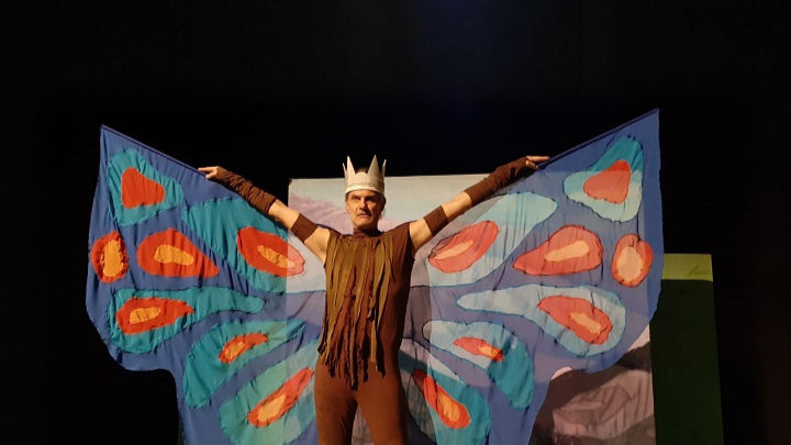 Nella foto Enrico Cavallero ha le braccia stese e alzate a mostrare le grandi ali colorate che ha applicate. lo sfondo è nero.