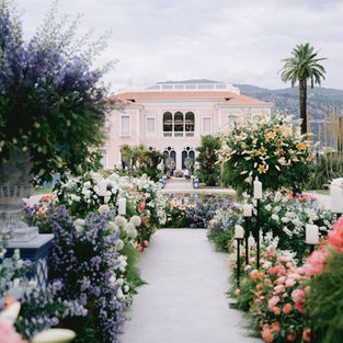 A gravel driveway with flower hedges on either side. In the background, a white villa with a tiled roof.