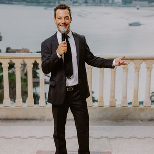 Jocelin Haas, in a black suit and white shirt, smiles with a microphone in front of his mouth. Behind Jocelin Haas, the sea and a boat.