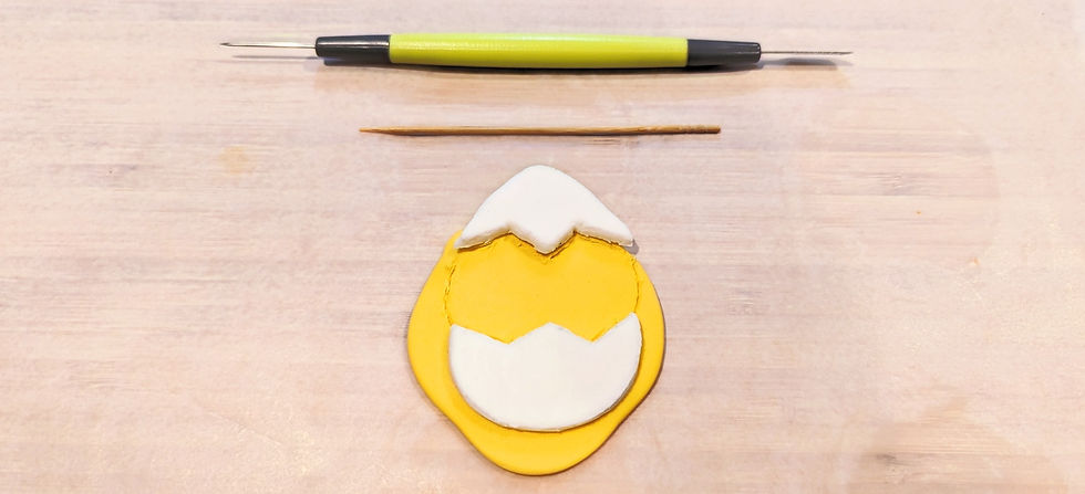 Two halfs of a fondant eggshell, sitting on top of a rolled out yellow fondant. Above the fondant a toothpick and fondant cutting tool is displayed.