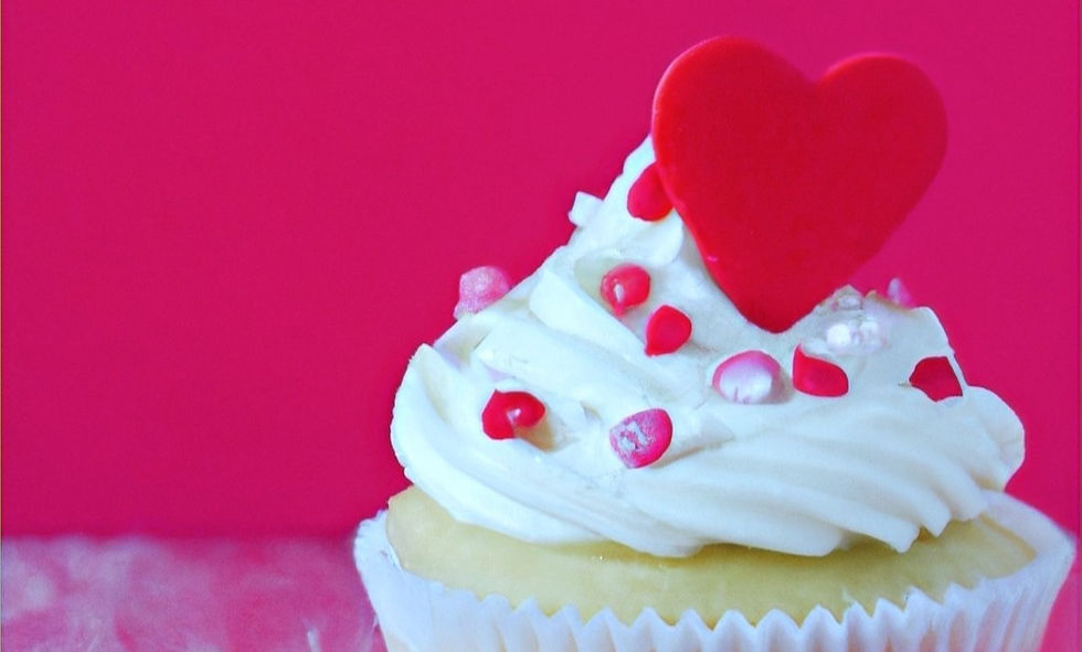 A vanilla cupcake with white frosting decorated with red and pink hearts, sitting in front of a pink background.