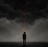 A lone silhouetted figure stands beneath black storm clouds with a thin red thread attached to his head, symbolizing ideological control and belief.