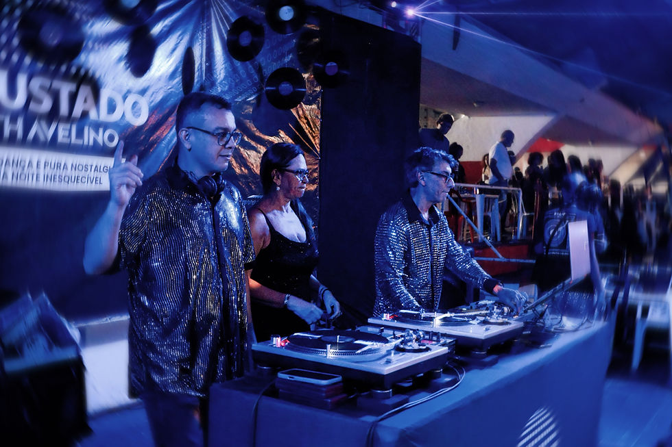 Três DJs em cabine com toca-discos sob luz azul; ao fundo, banner com texto desfocado e público em festa.