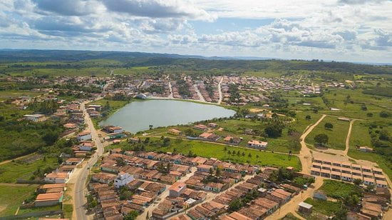 Vista aérea de cidade pequena com lago central, casas e ruas, cercada por campos verdes sob céu nublado.