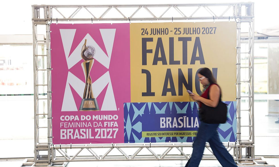 Mulher caminha lendo celular diante de cartaz da Copa do Mundo Feminina FIFA Brasil 2027, com texto FALTA 1 ANO e BRASÍLIA.